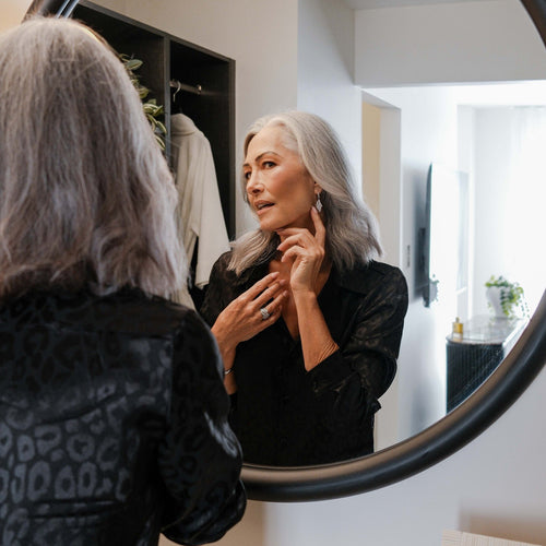 Women with long grey hair admiring diamond drop earrings in a round bedroom mirror.
