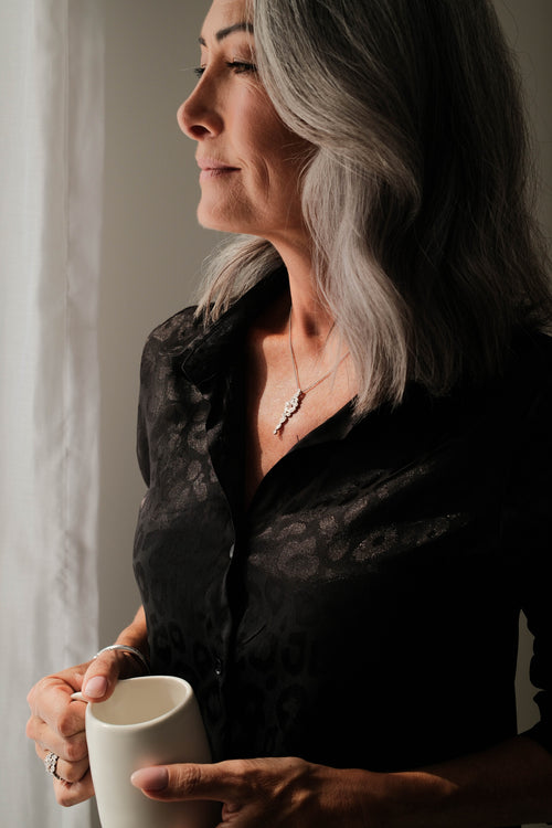 Woman with gray hair holding a white mug, wearing a black shirt and a diamond pendant necklace