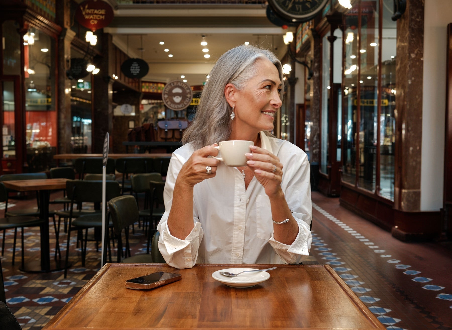 A woman wearing diamond earrings, cuff bracelet and diamond rings having a coffee at a table inside Sydney's Strand Arcade