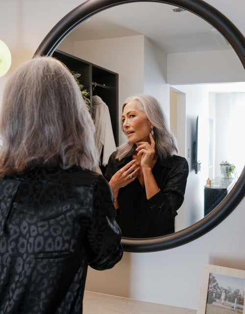 Women with long grey hair admiring diamond drop earrings in a round bedroom mirror.