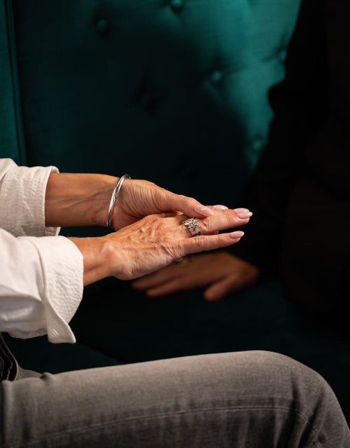 A close-up lifestyle shot of a person's hands resting on their lap, wearing a silver bangle bracelet and a silver ring with a dark stone, seated against a dark green tufted velvet background.