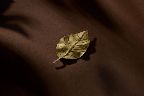 Gold single leaf brooch with sparkling crystal accents on a dark brown background.