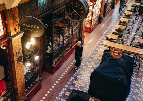 View from above of a woman walking on a ornately tiled floor. In the foreground is a shop sign that reads Mondial By Nadia Neuman and behind that a large vintage clock with roman numerals