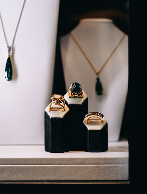 Three gold rings displayed on black and white jewelry stands, with a white bust displaying a gold necklace with a dark teardrop pendant in the background.