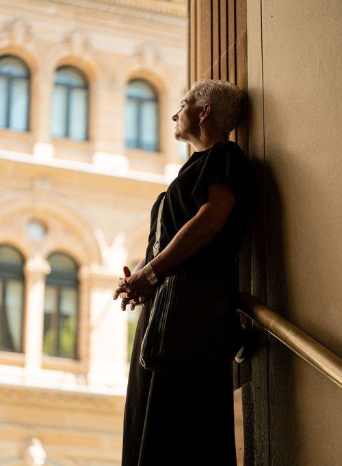 A woman with short blonde hair wearing a black dress stands in profile, leaning against a wall in a sunlit interior next to a window, with a blurred classical building facade in the background.