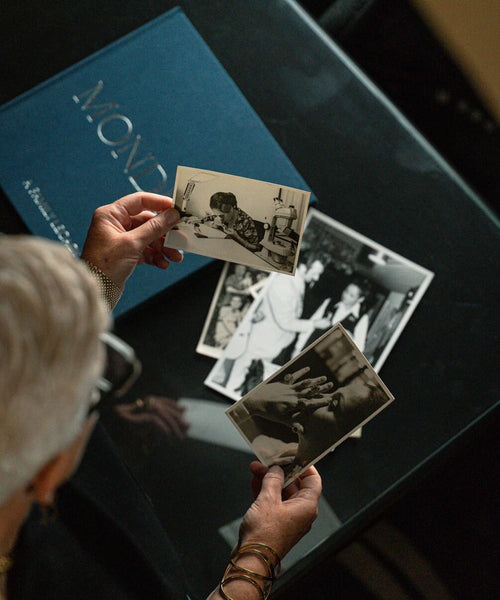 A person holds two vintage black-and-white photographs over a dark table, with a blue book or album cover partially visible in the background.