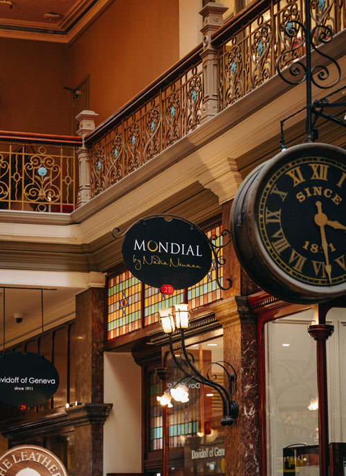 A vintage-style hanging wall clock with Roman numerals and the text 'SINCE 1816' inside The Stand Arcade, a grand indoor shopping mall in Sydney. A black oval sign reading 'MONDIAL by Nadia Neuman' is visible below, along with ornate balconies, warm wood-toned architecture, and other storefront signage.