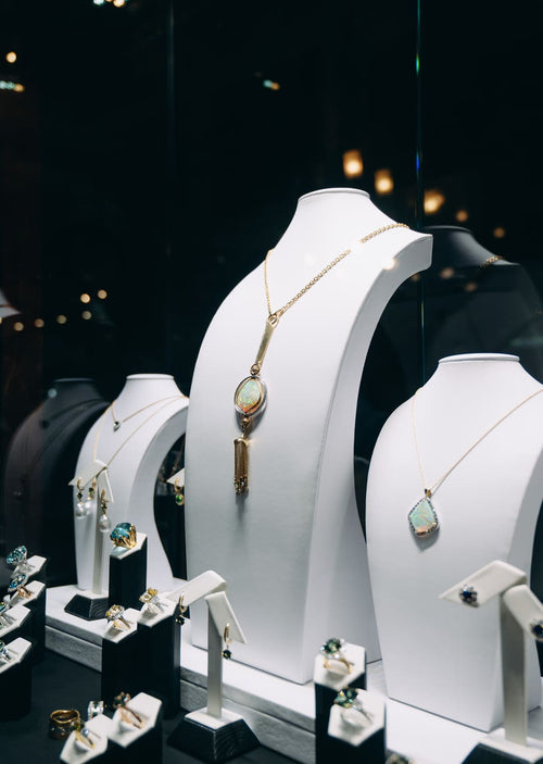 Gold jewelry display busts in a boutique setting, featuring a gold pendant necklace with a light green opal and a gold key-shaped pendant, alongside other necklaces and rings on black display stands.